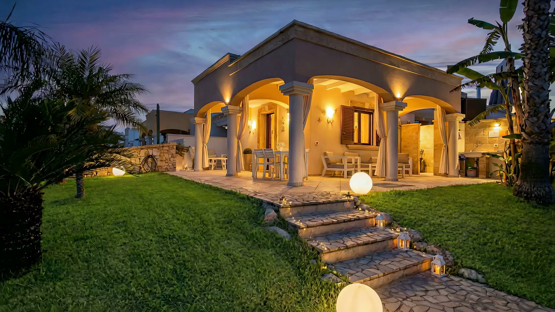 Casa Pescoluse 8 al tramonto: la veranda ad archi illuminata di luce calda, le lanterne sulla scalinata in pietra e le sfere luminose sul prato