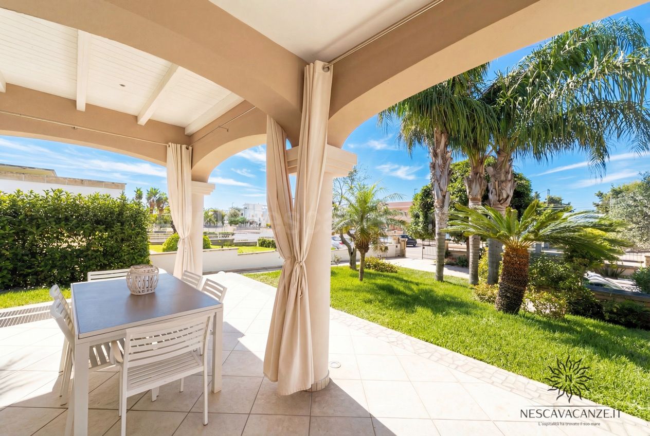 Casa Pescoluse 8 - Veranda con tavolo da pranzo, tende chiare e vista sul giardino con palme