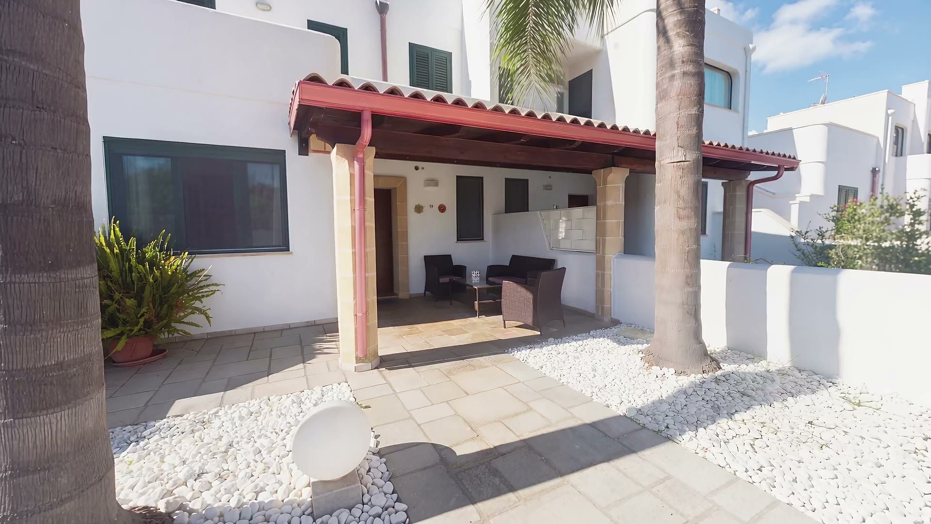 Casa Sabbia d'Oro oltre il cancello: il cortile con i ciottoli bianchi e le palme, il salottino in rattan sotto la tettoia in coppi e la porta d'ingresso in legno