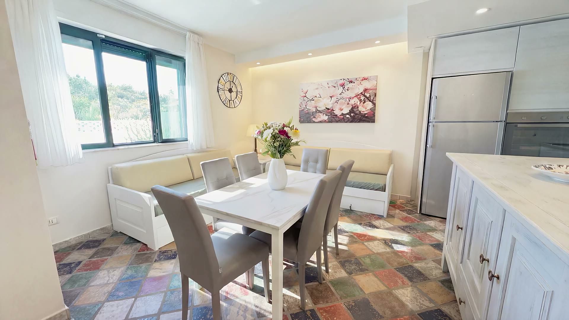 Il soggiorno di Casa Sabbia d'Oro: i divani bianchi sotto la finestra, il tavolo da pranzo, la TV e il pavimento in cotto policromo