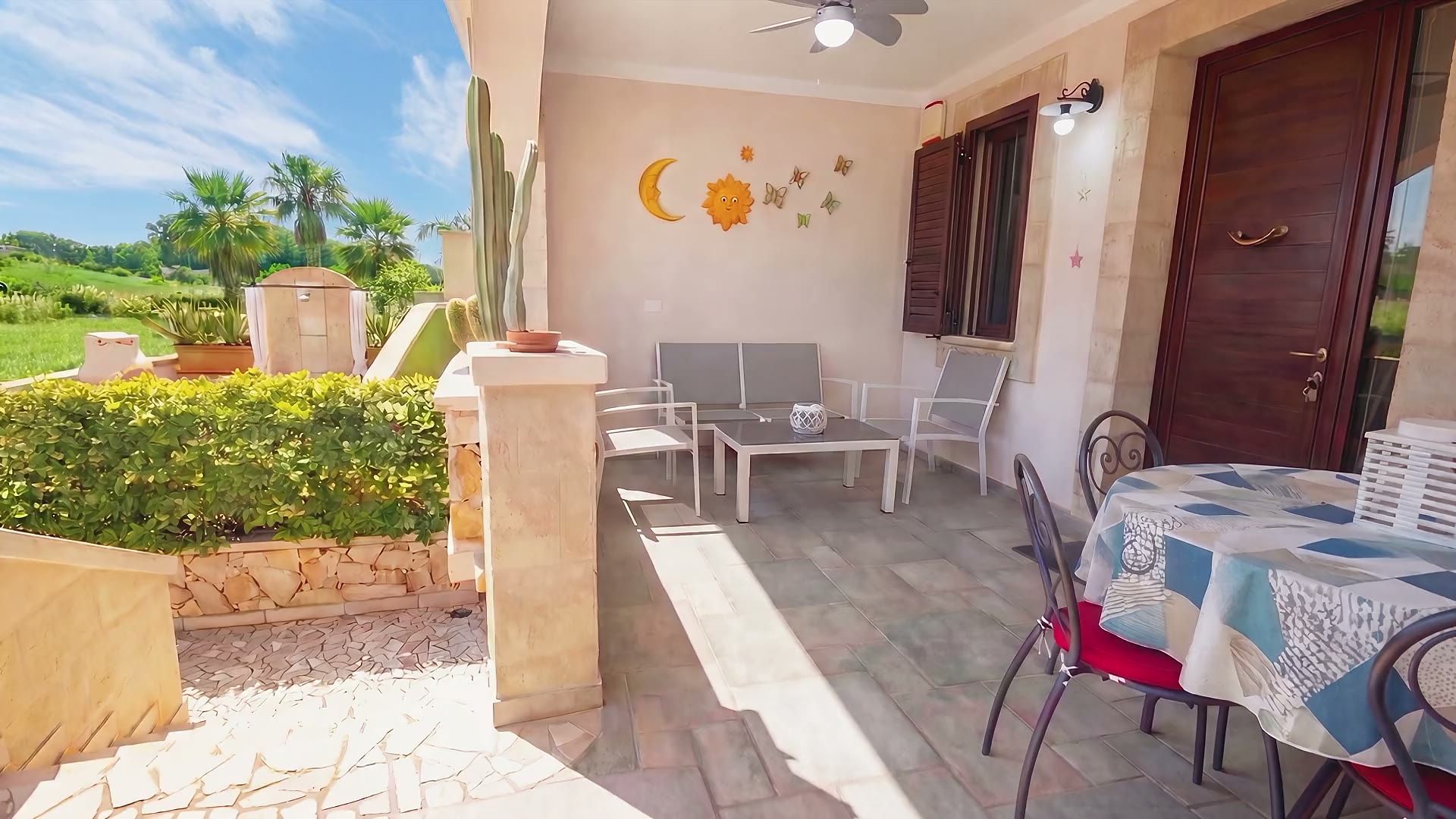 La veranda di Casa Pescoluse 4 con il divanetto grigio, il tavolo tondo con la tovaglia a patchwork e la porta d'ingresso in legno