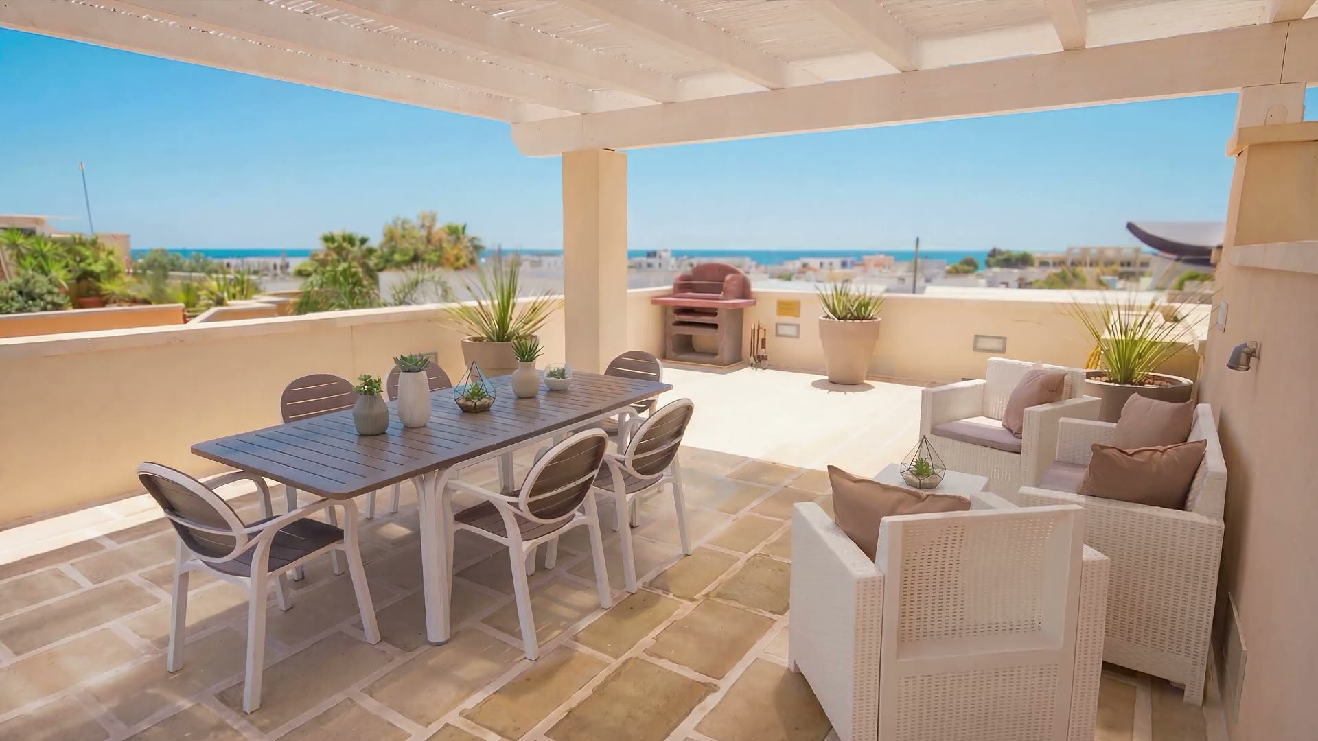 La terrazza sul tetto di Casa Giglio sotto la pergola bianca, con il tavolo da pranzo, i divani in rattan, il barbecue in muratura e il mare all'orizzonte