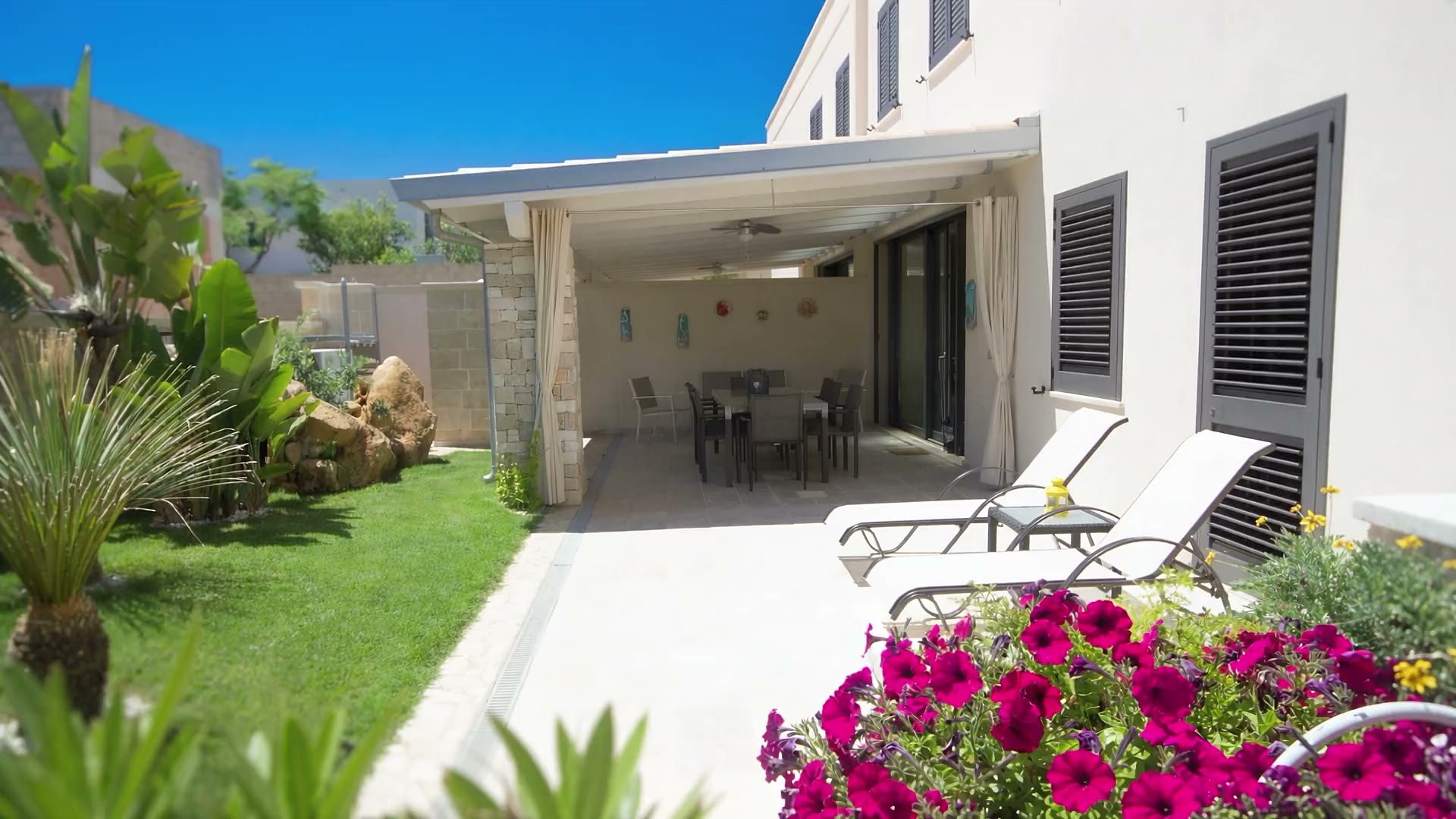 Il giardino privato di Casa Dune con il prato, le palme, i lettini prendisole sulla pietra chiara e la veranda in legno addossata alla casa