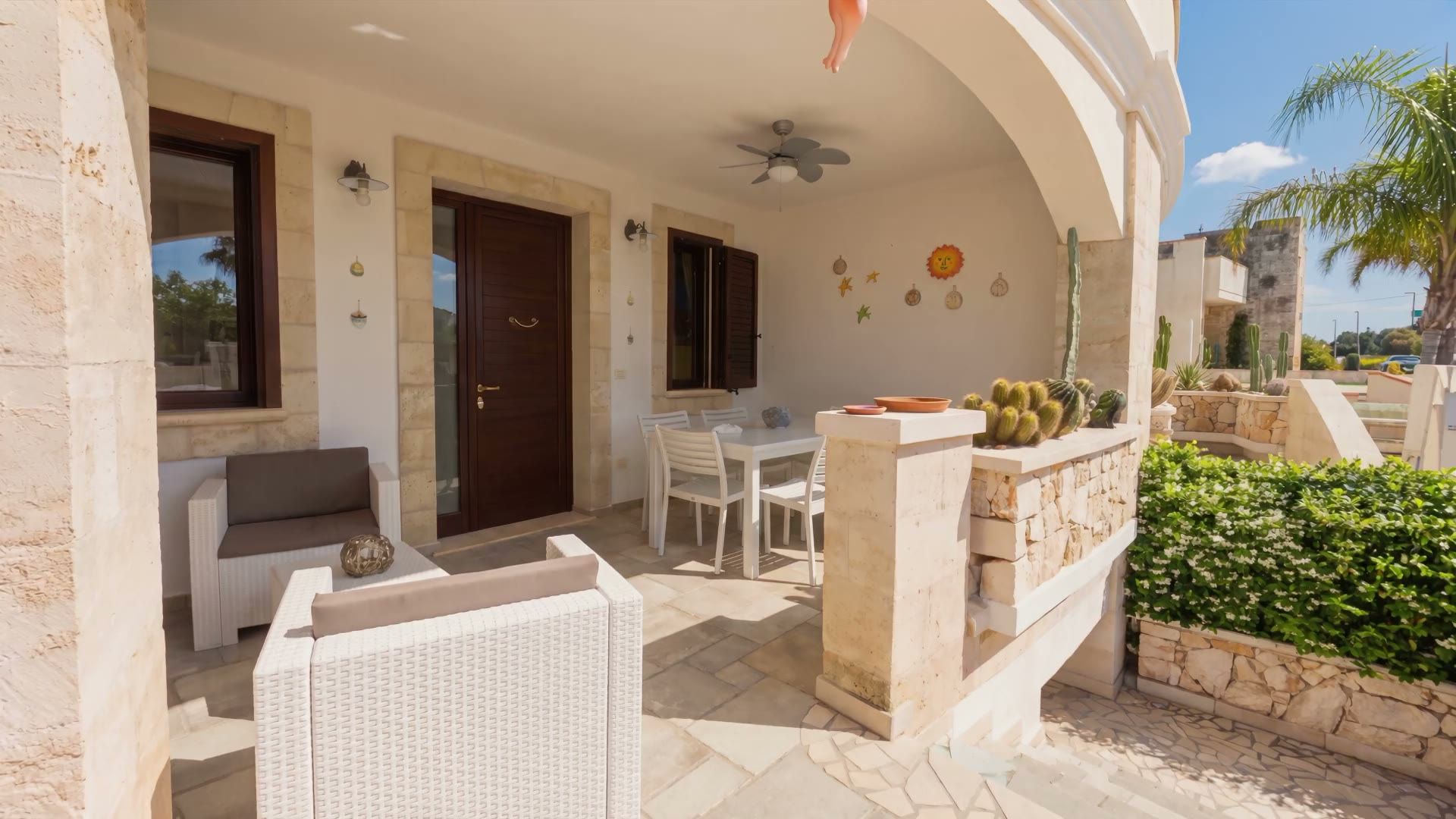 La veranda coperta di Casa Pescoluse 5 con il tavolo da sei, le poltrone in rattan, il ventilatore a pale e la porta d’ingresso in legno