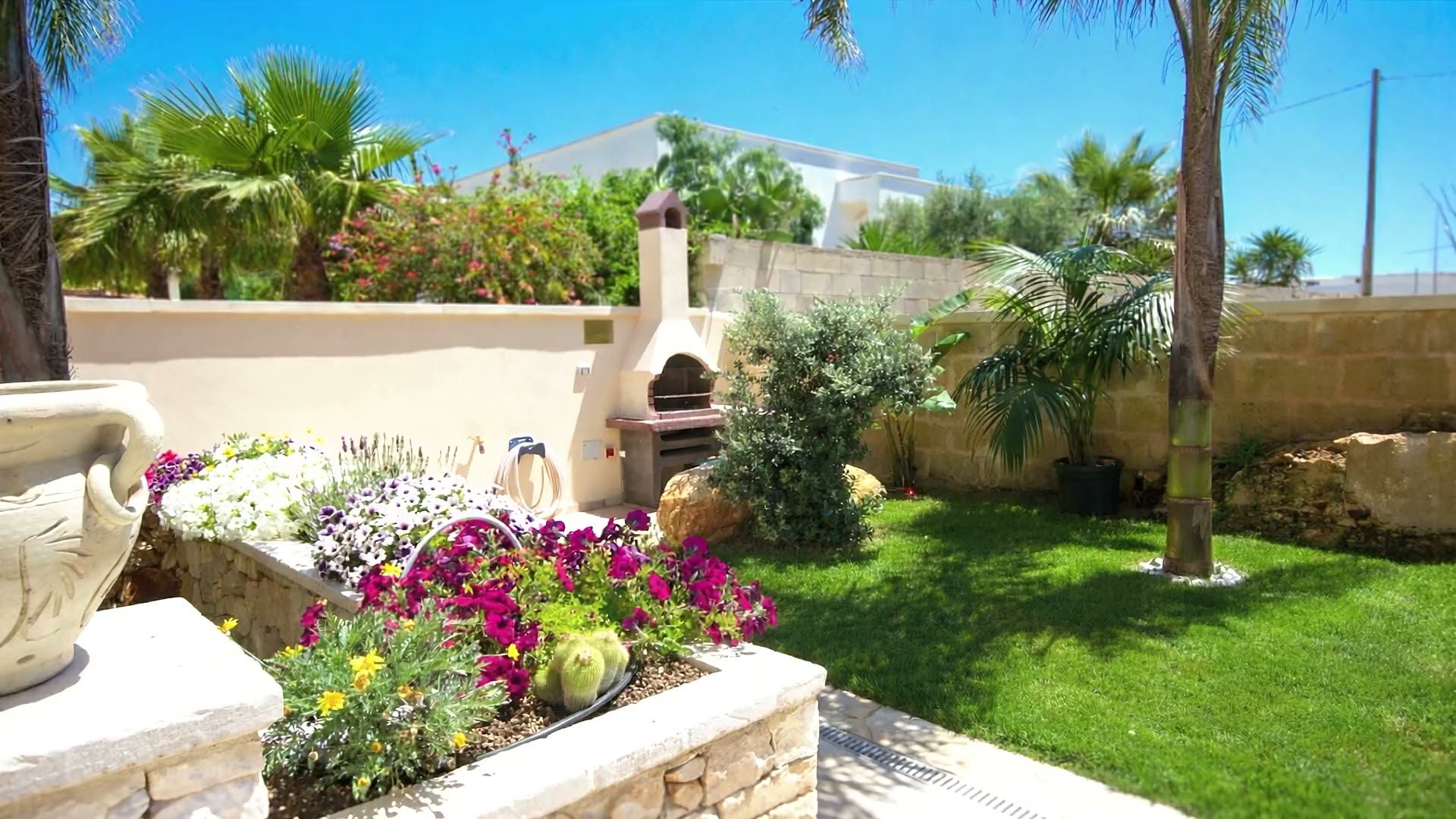 Il prato di Casa Dune con le palme, il vaso in terracotta, il barbecue in muratura e il muro di cinta con le case vicine oltre