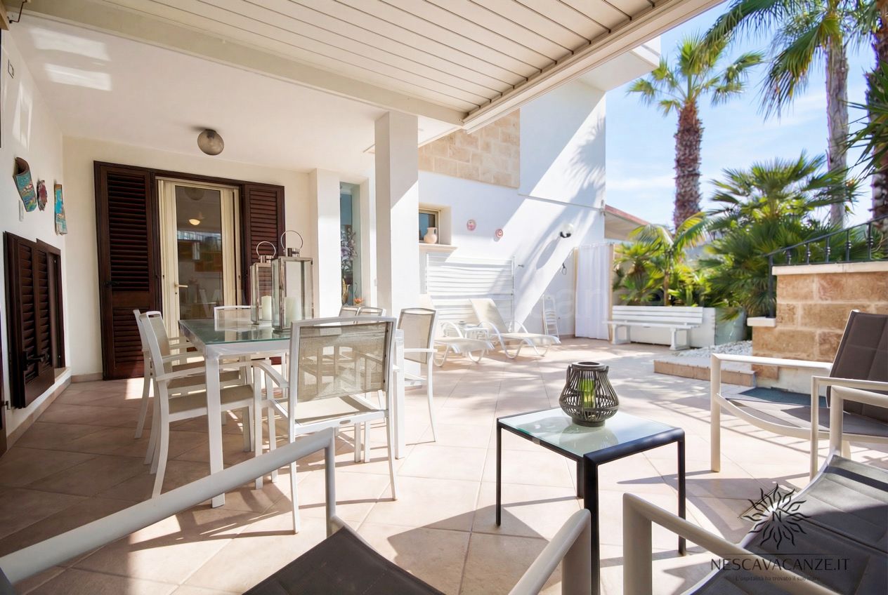 Casa Pescoluse 2 - patio coperto con tavolo, lettini prendisole e palme sullo sfondo