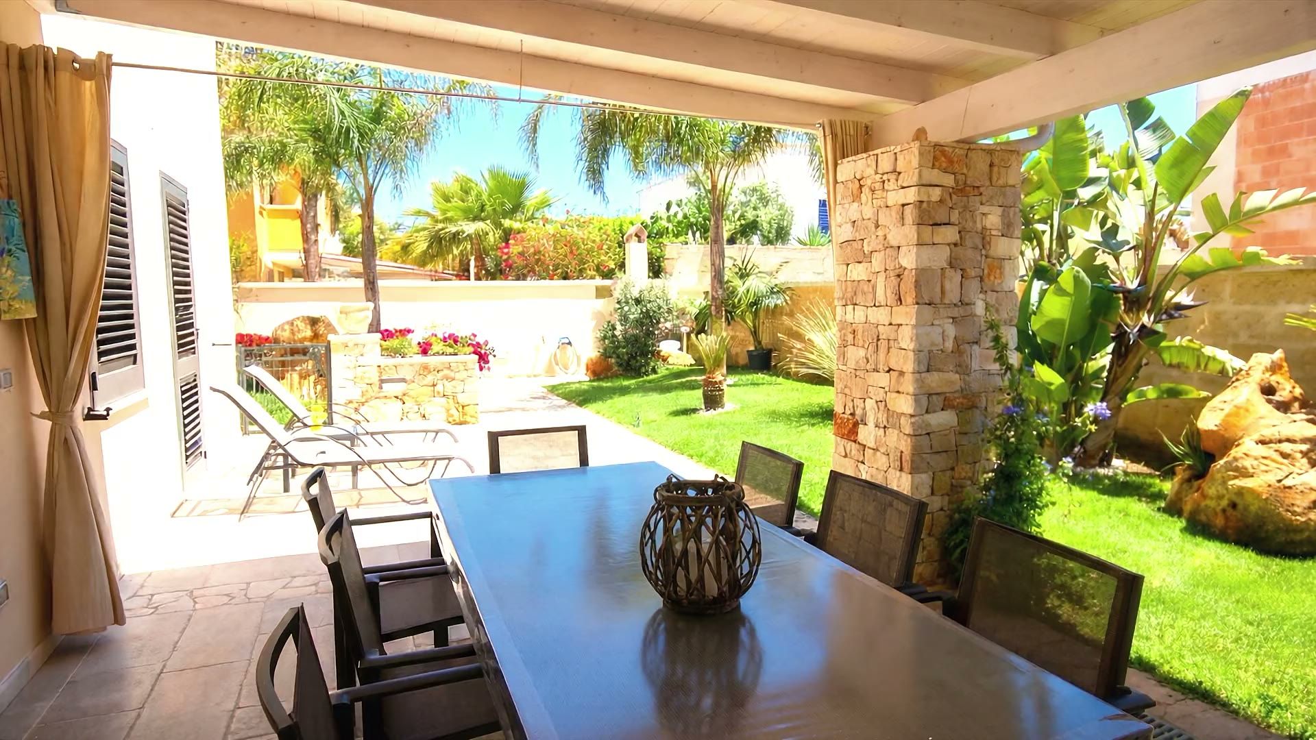 La grande veranda in legno di Casa Dune con il tavolo da pranzo esterno, i divani in rattan e la vetrata scorrevole sul soggiorno