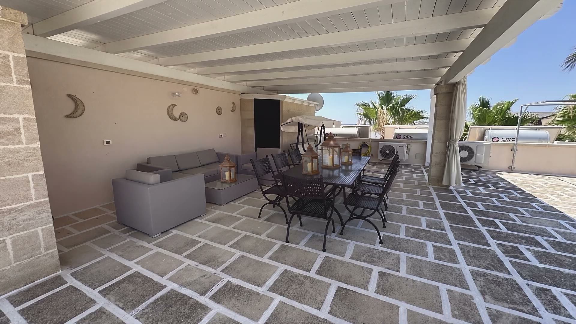 Sotto la tettoia del terrazzo di Casa Pescoluse 4: il lungo tavolo in ferro battuto, i divanetti grigi e il dondolo