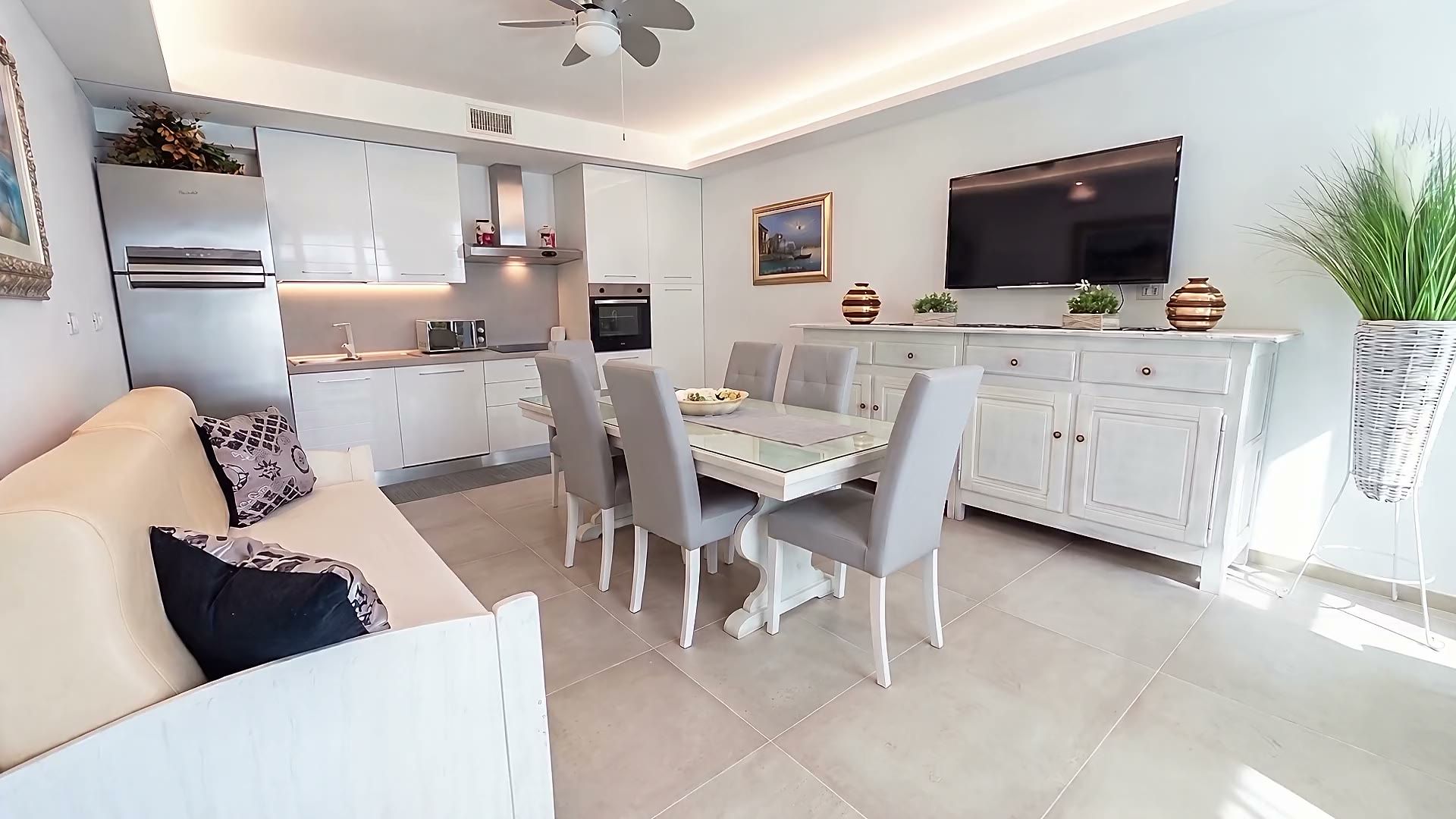 Il soggiorno di Casa Pescoluse 2 con il divano, il tavolo da pranzo con le sedie grigie e la cucina bianca attrezzata