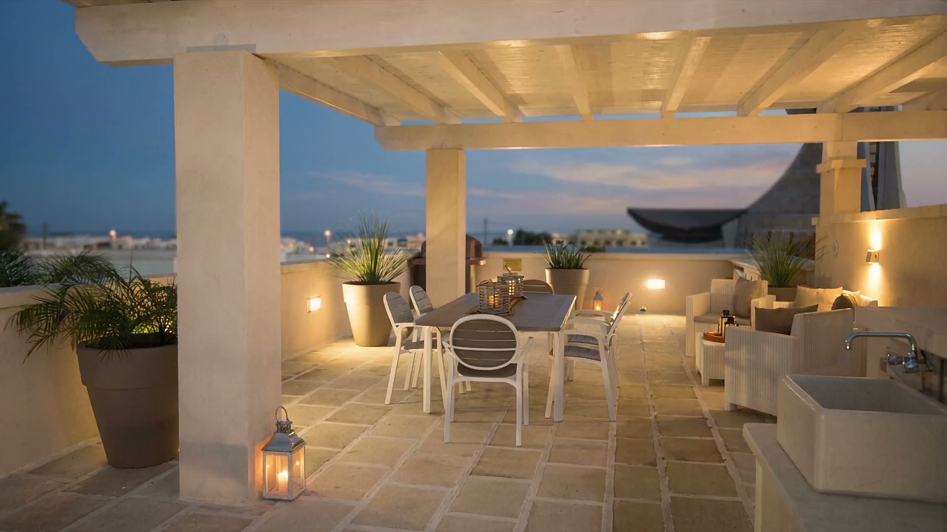 La terrazza di Casa Giglio al crepuscolo, con le luci accese sotto la pergola, le lanterne sul tavolo e il cielo blu sui tetti