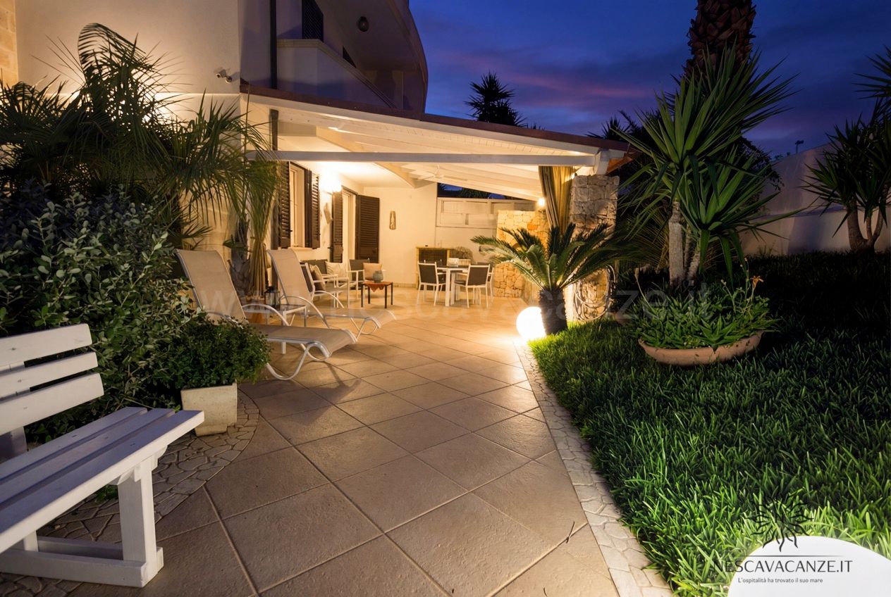Casa Pescoluse 3 - patio illuminato al crepuscolo con lettini, panca bianca e tavolo sotto la veranda nel giardino