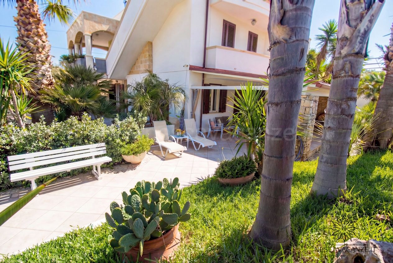 Casa Pescoluse 3 - giardino soleggiato con lettini prendisole, panca bianca e piante grasse davanti alla facciata