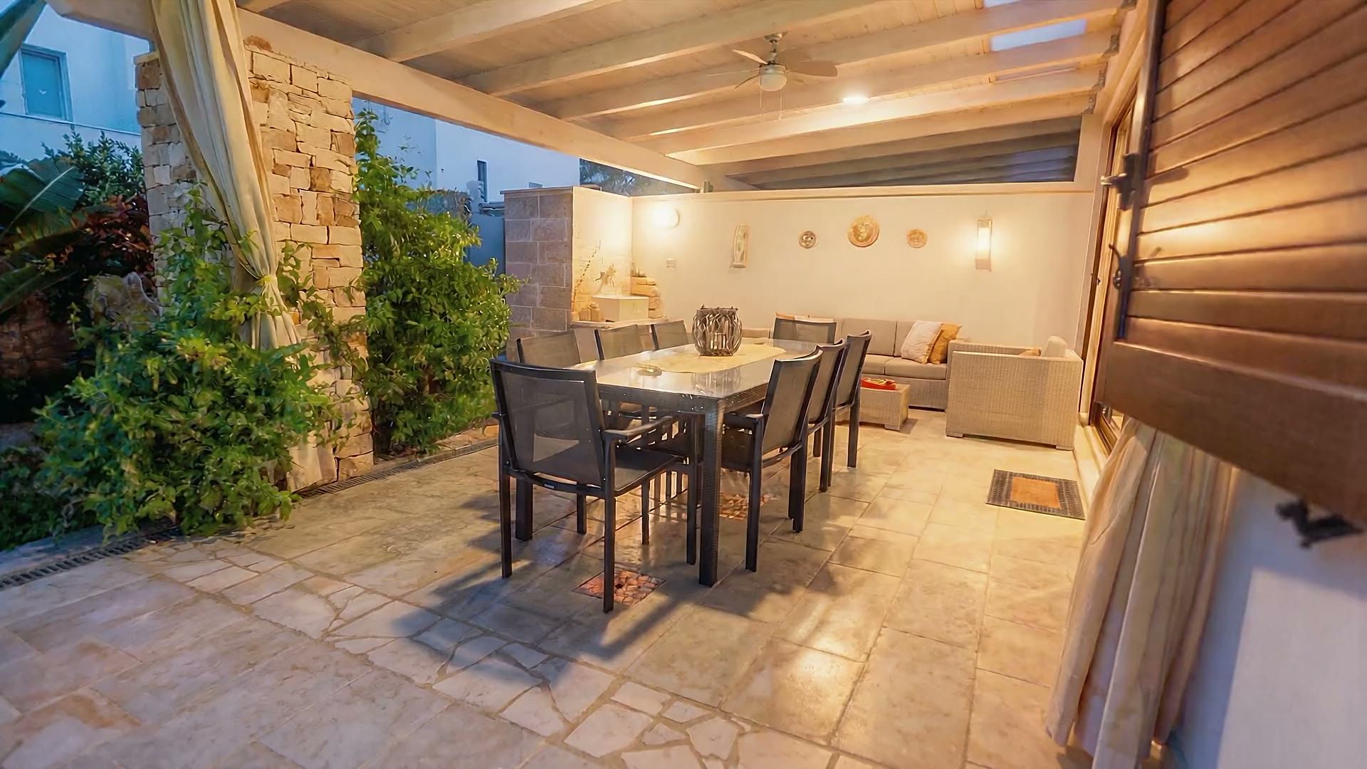La veranda di Casa Dune illuminata la sera, con il tavolo da pranzo, la lanterna, i divani e l'ultimo blu del cielo oltre la tettoia