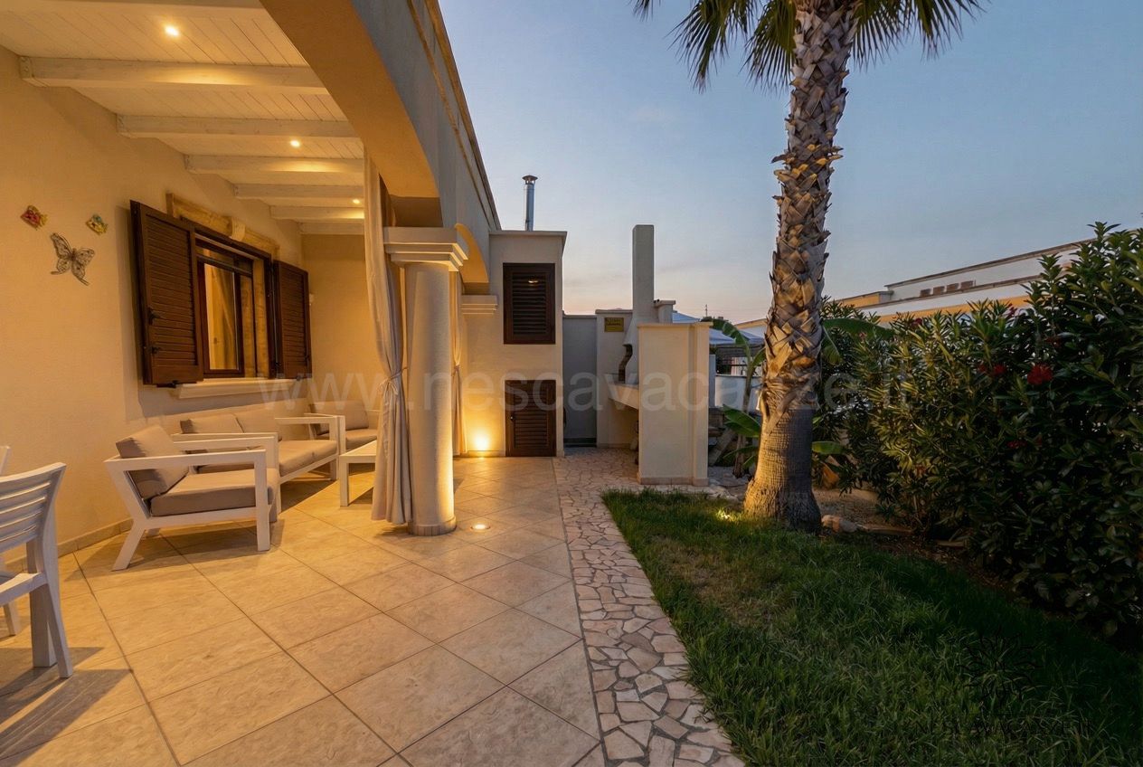 Casa Pescoluse 8 - Veranda laterale al tramonto con divanetti, zona barbecue e palma illuminata