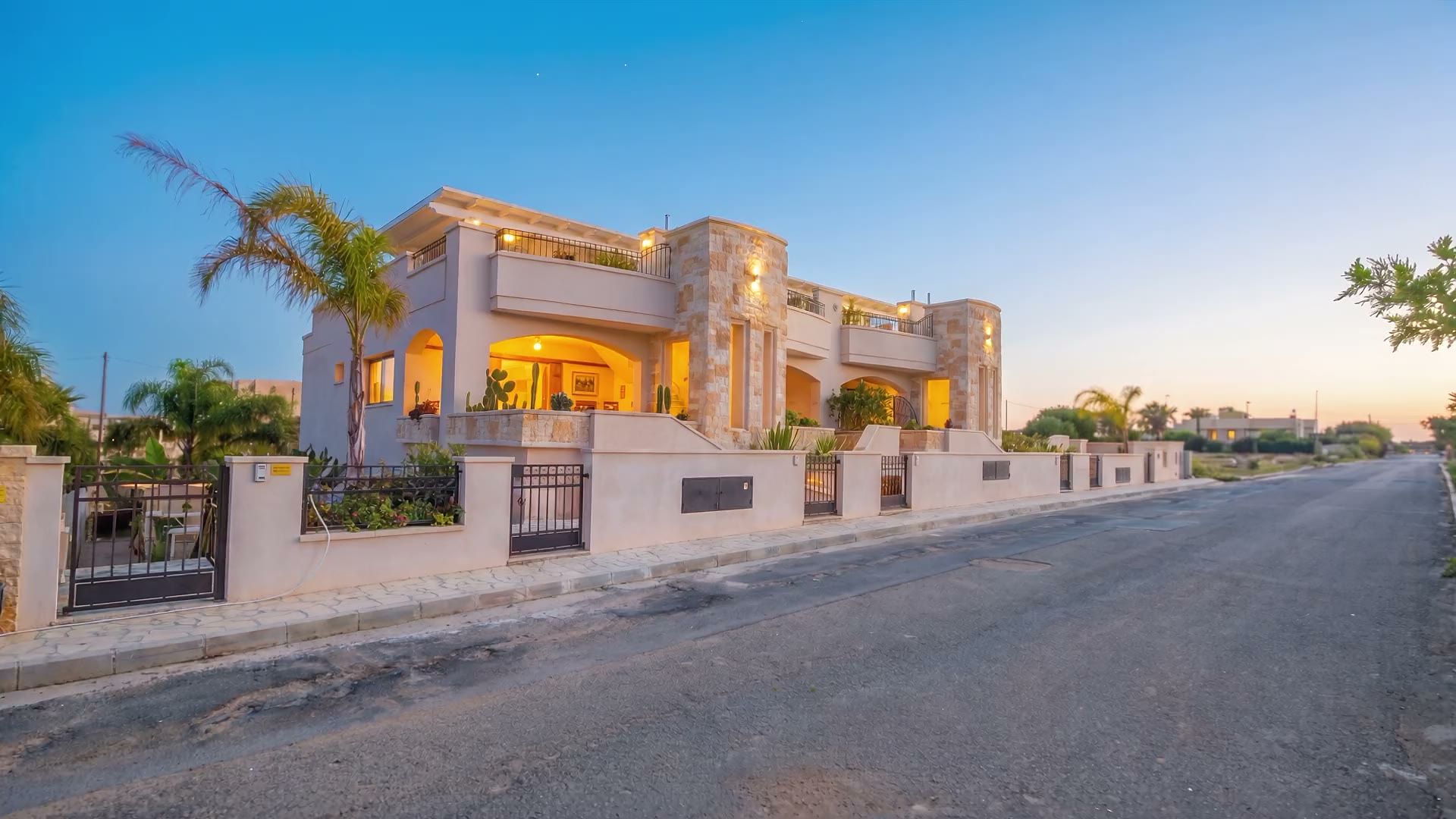 La via davanti a Casa Dune al crepuscolo, con la fila delle villette in pietra chiara, le luci accese e le palme contro il cielo blu della sera