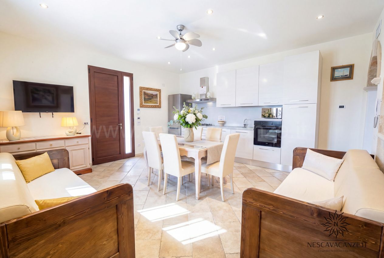 Casa Pescoluse 8 - Zona giorno luminosa con cucina bianca, tavolo, divani e televisore a parete