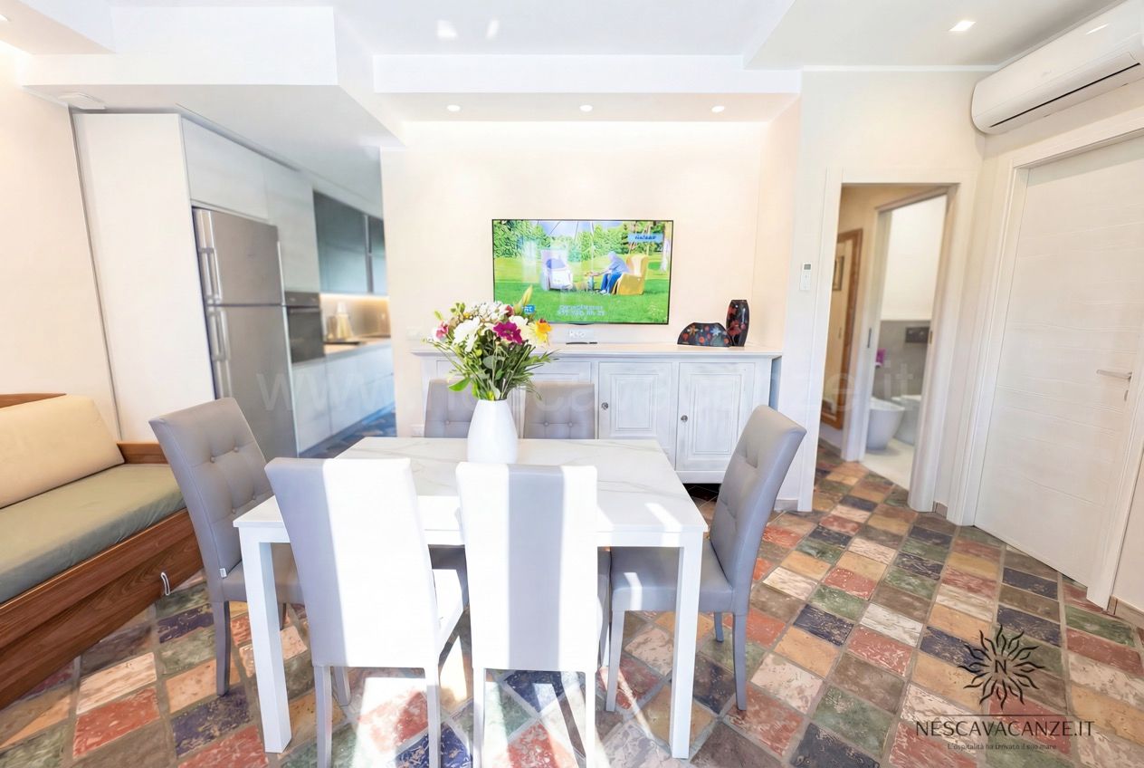 Casa Sabbia Oro - zona pranzo con tavolo bianco, TV, cucina sullo sfondo e pavimento colorato