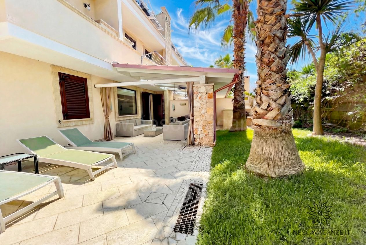 Casa Pescoluse 7 - giardino con lettini verdi, palma e patio coperto con divani sullo sfondo