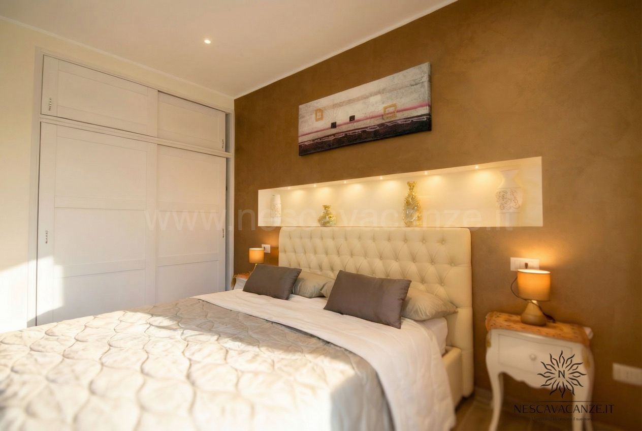 Casa Dune - camera matrimoniale con letto, testiera imbottita, armadio bianco, parete marrone e luci decorative nello scaffal
