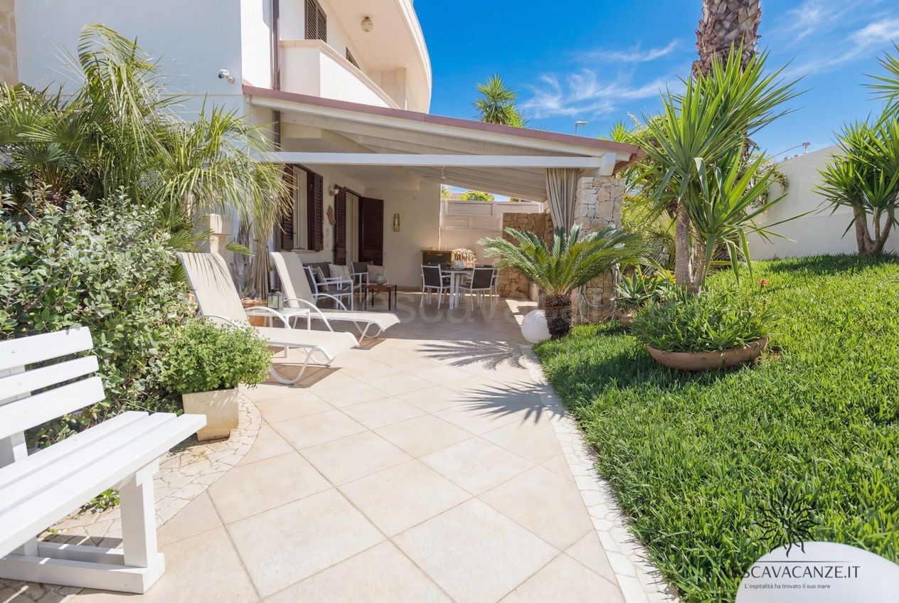 Casa Pescoluse 3 - patio esterno con piastrelle chiare, lettini, tavolo da pranzo sotto pergolato e prato verde con palme