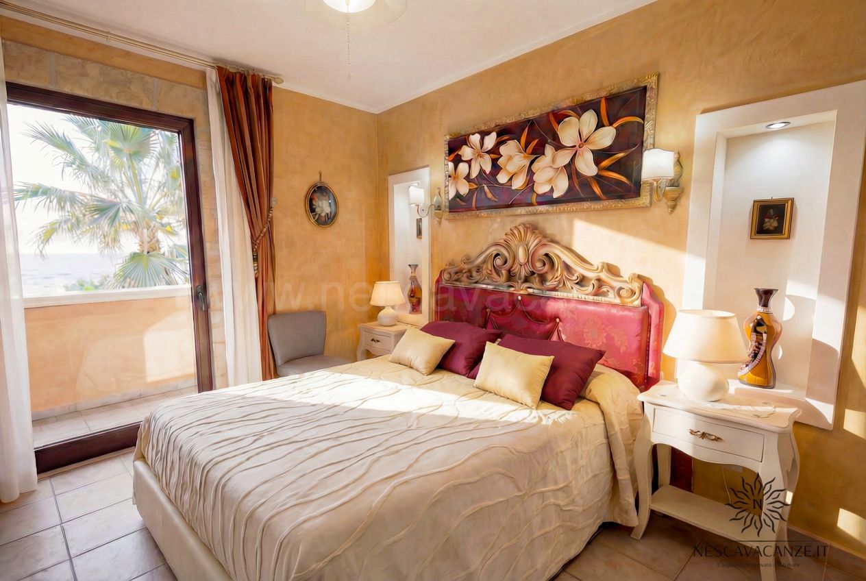 Casa Pescoluse 5 - camera matrimoniale con testiera rossa, quadro floreale e balcone con vista palme