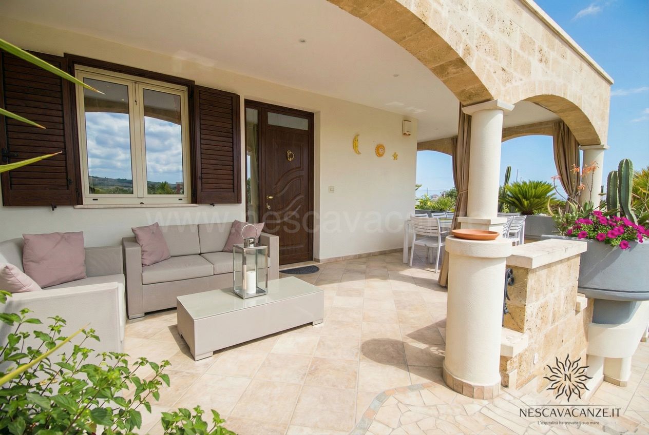 Casa Pescoluse 1 - veranda coperta con divani, cuscini, tavolino, lanterna e tavolo da pranzo sullo sfondo
