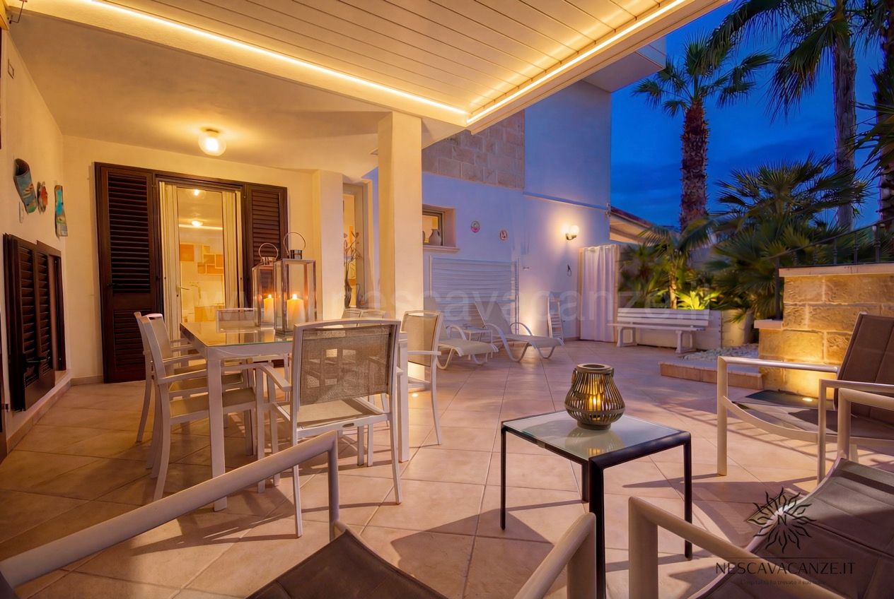 Casa Pescoluse 2 - patio coperto al tramonto con tavolo, lanterne, lettini e palme illuminate