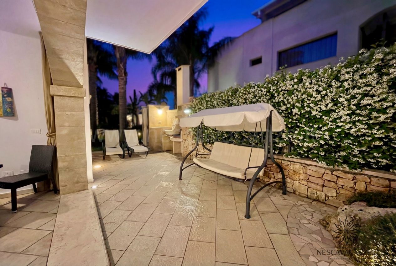 Casa Pescoluse 6 - patio illuminato al crepuscolo con dondolo, lettini e siepe di gelsomino
