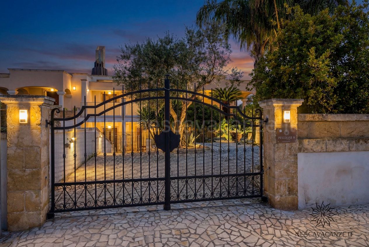 Casa Pescoluse 9 - cancello in ferro battuto al tramonto con villa illuminata e cielo rosato