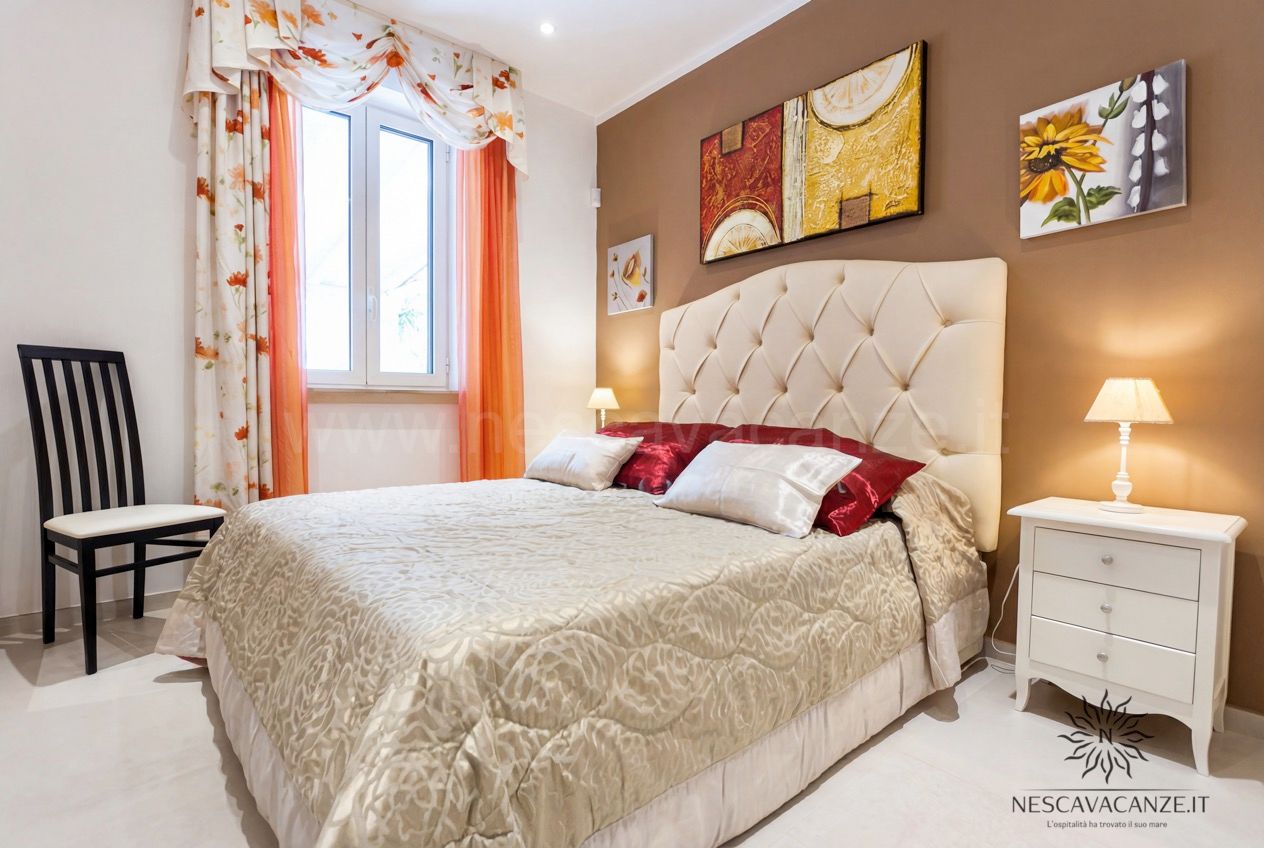 Casa Pescoluse 3 - camera matrimoniale con testiera capitonné beige, comodini bianchi e tende arancioni fiorite