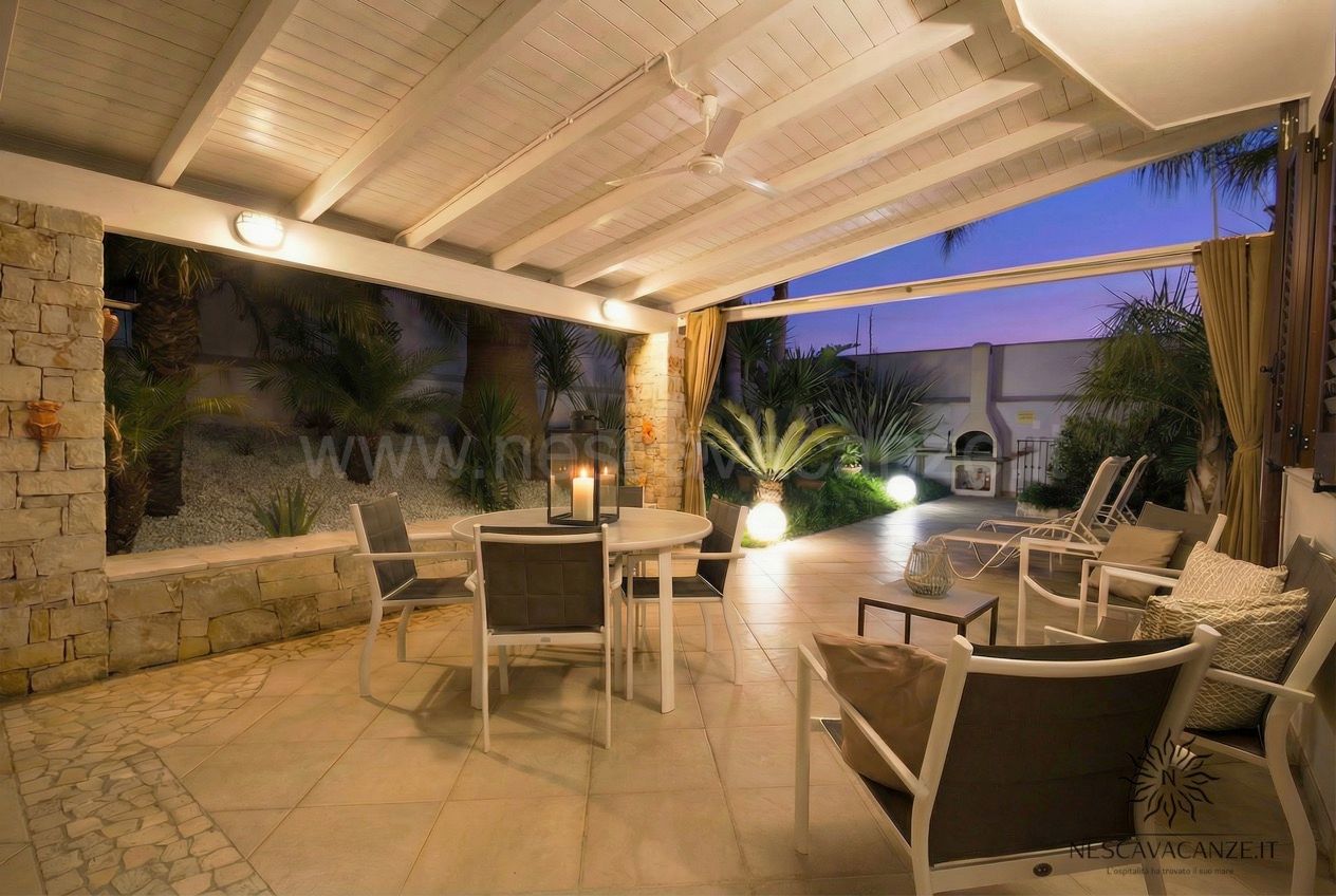 Casa Pescoluse 3 - patio coperto illuminato al crepuscolo con tavolo rotondo, sedie e poltrone nel giardino