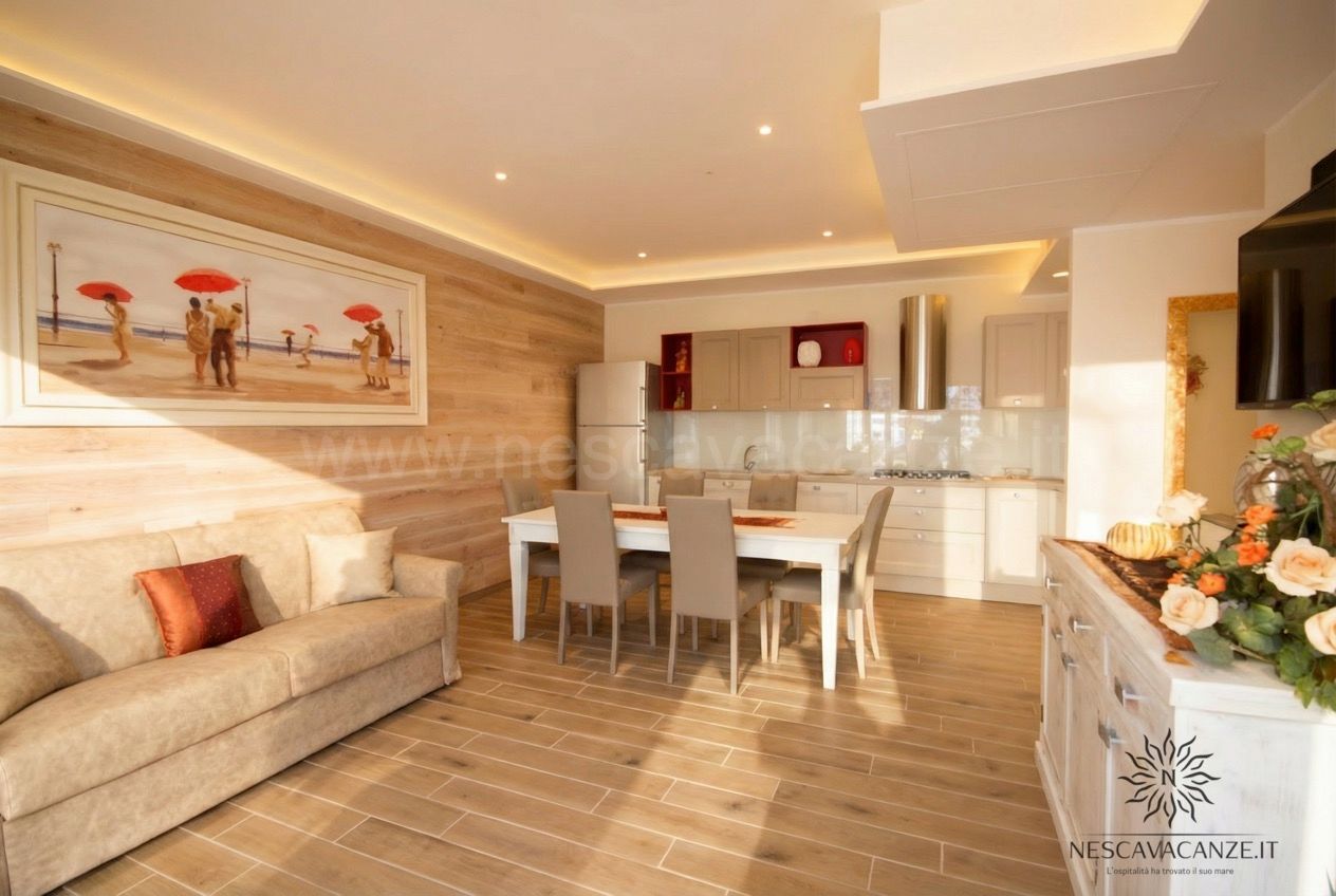 Casa Dune - soggiorno con divano beige, tavolo da pranzo bianco, cucina a vista chiara e quadro con persone sulla spiaggia