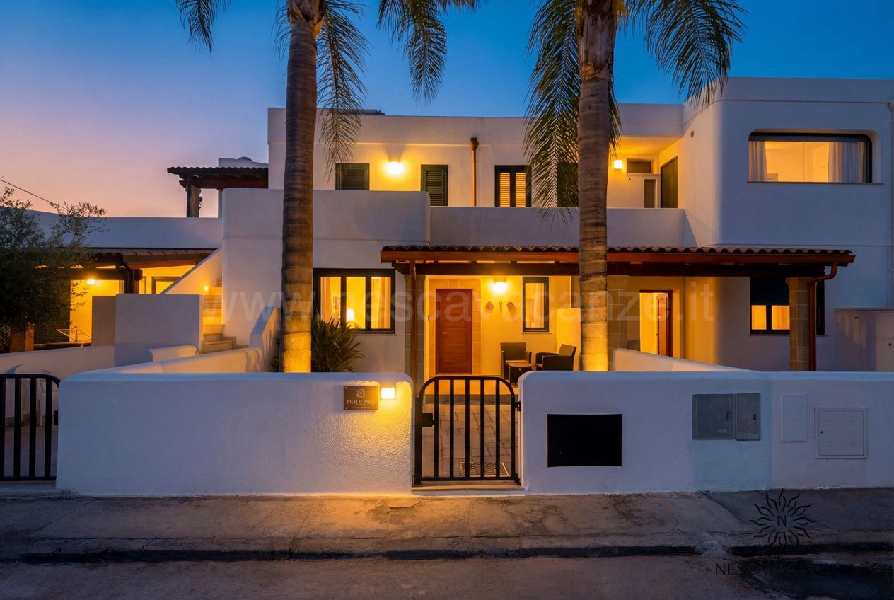 Casa Sabbia Oro - facciata illuminata al tramonto con palme, veranda e cielo crepuscolare