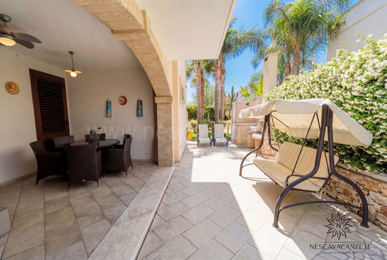 Casa Pescoluse 6 - veranda coperta con salotto in rattan, dondolo e palme sul patio soleggiato