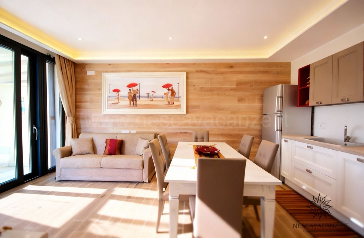 Casa Dune - soggiorno con divano beige, tavolo da pranzo bianco, cucina a vista con pensili grigi e frigorifero, pavimento in