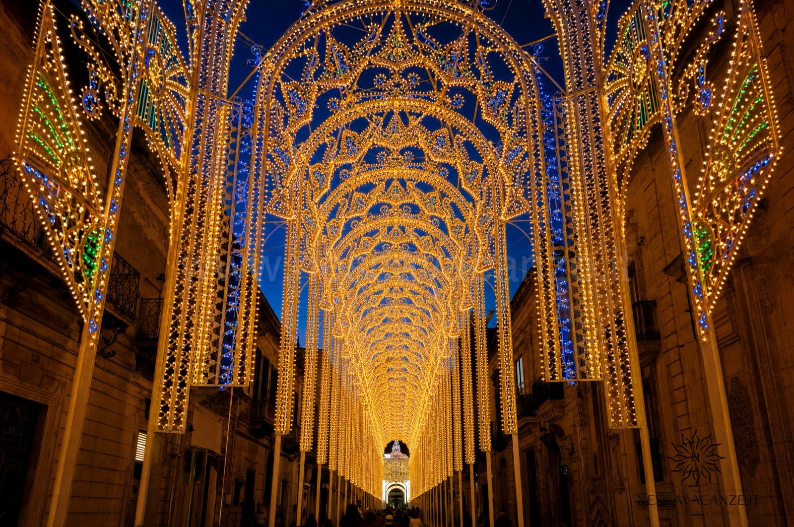 Luminarie salentine sopra la via di un paese in festa: archi di legno illuminati da migliaia di lampadine, come per San Nicola a Salve