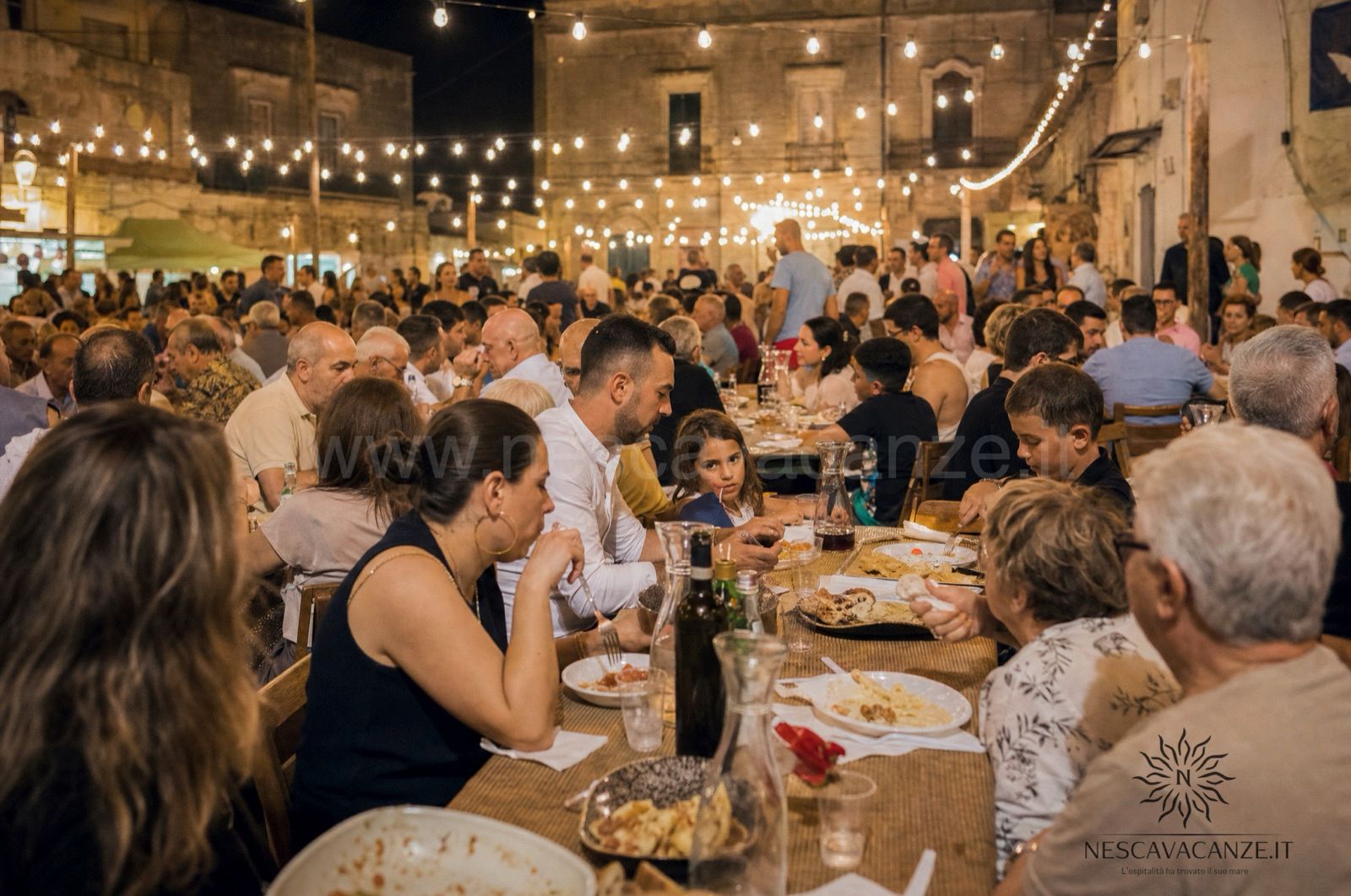 Lunga tavolata conviviale all'aperto durante una sagra di paese salentina, illuminata di sera