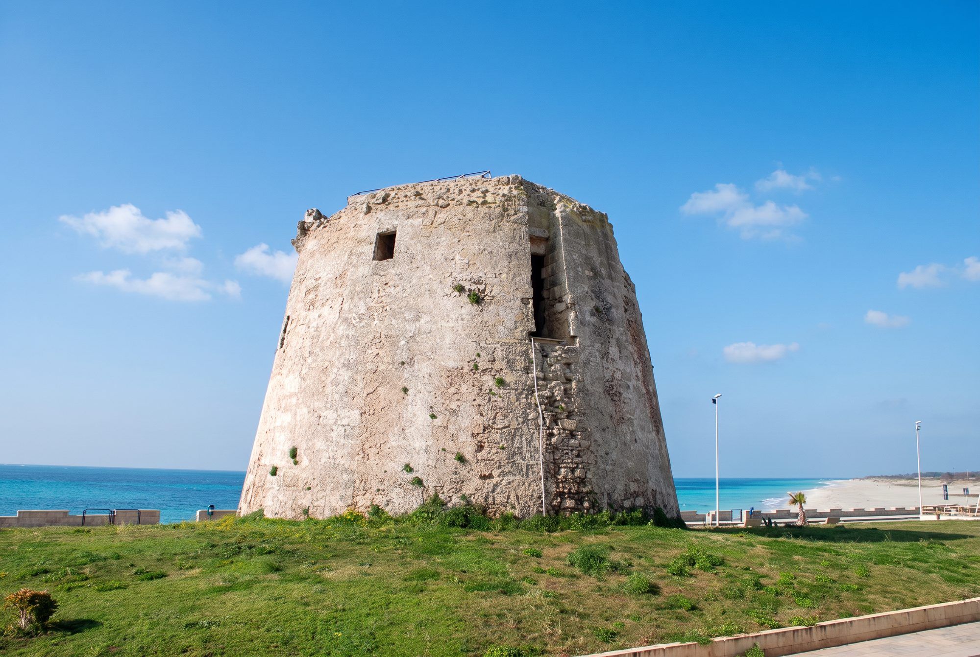 La torre tronca di Torre Mozza sul prato, con il mare turchese sullo sfondo