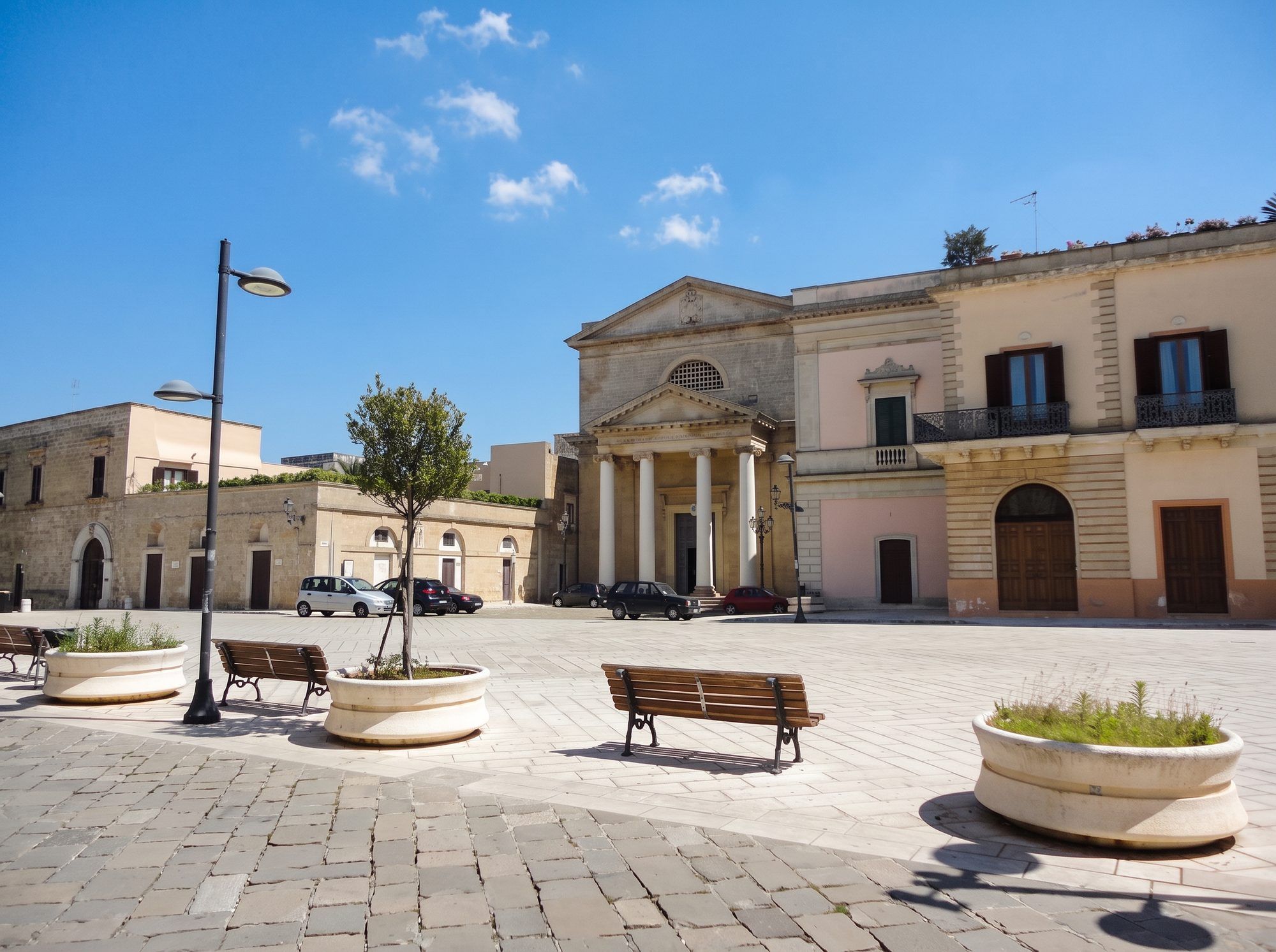 La piazza di Ugento con le panchine, i lampioni e la facciata a colonne di una chiesa