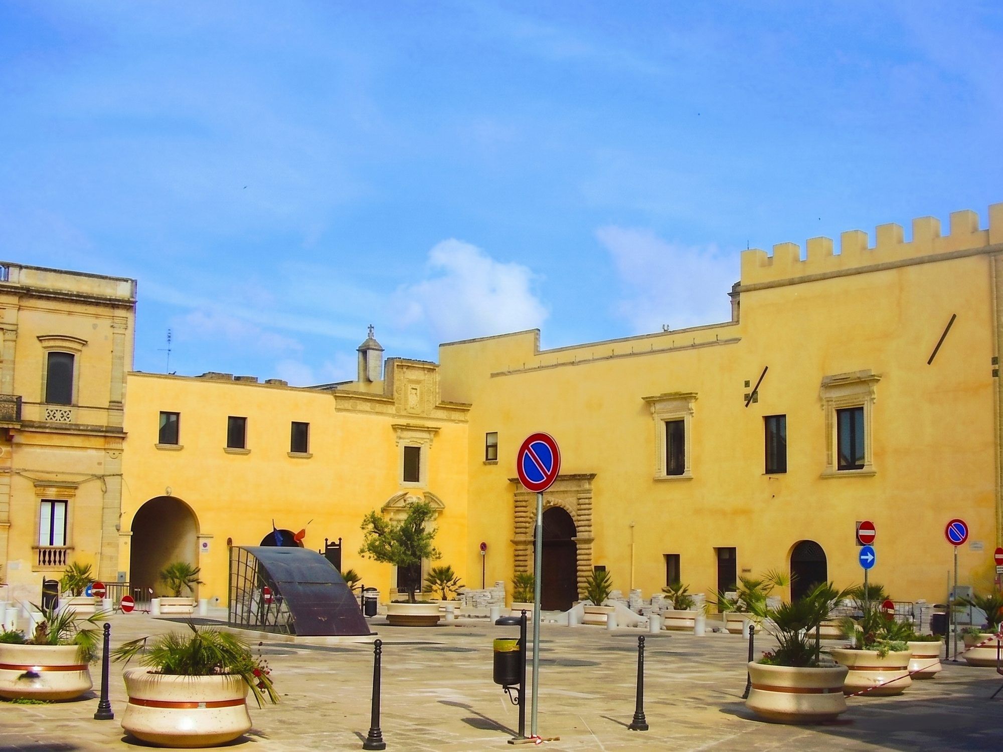 Piazza del Popolo a Presicce con il Palazzo Ducale giallo e merlato, i vasi di palme e il cielo azzurro