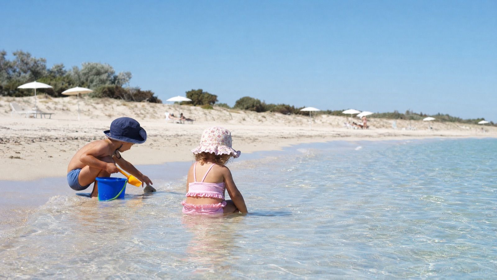 Un bambino e una bambina con il cappellino giocano con secchiello e paletta sulla battigia, tra dune basse e acqua trasparente alta pochi centimetri