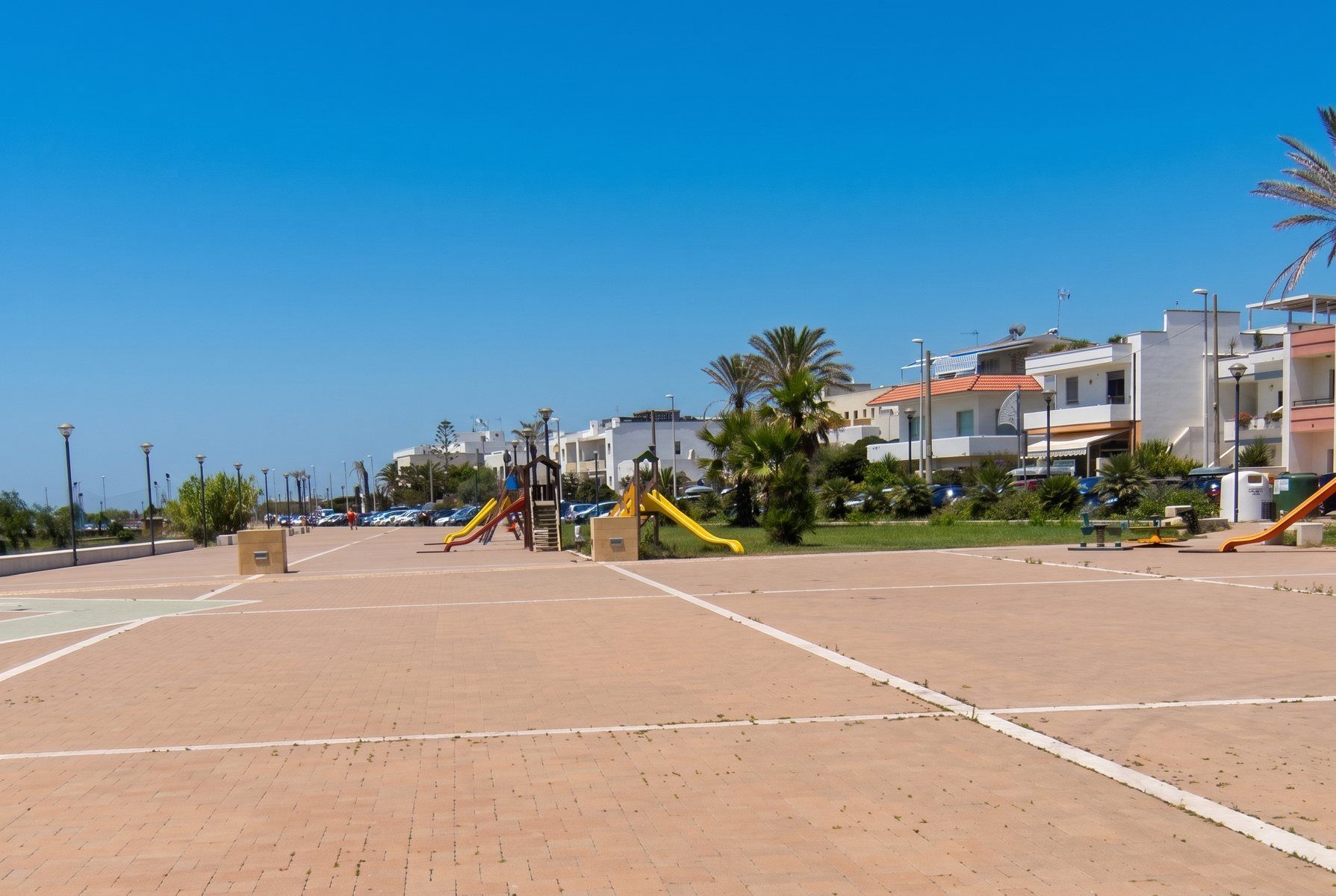Piazza sul lungomare di Pescoluse con palme, giochi per bambini e case basse