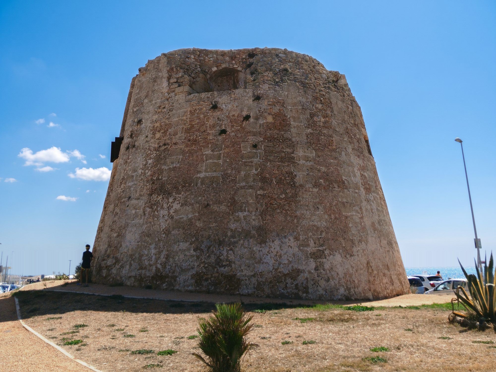 La torre tronca di Torre Mozza su un prato, con le auto parcheggiate e il mare sullo sfondo a destra