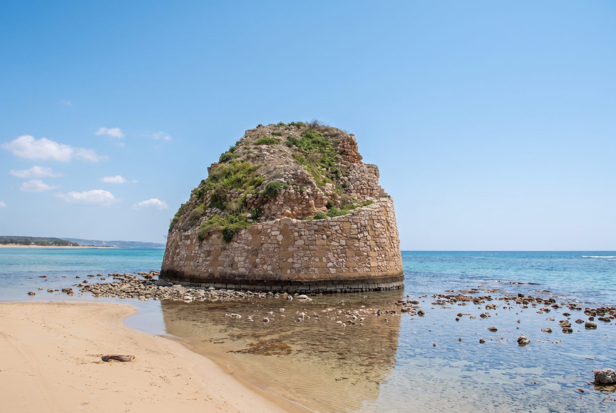 La torre di Torre Pali riflessa nell'acqua bassa, con la riva di sabbia e la costa sullo sfondo