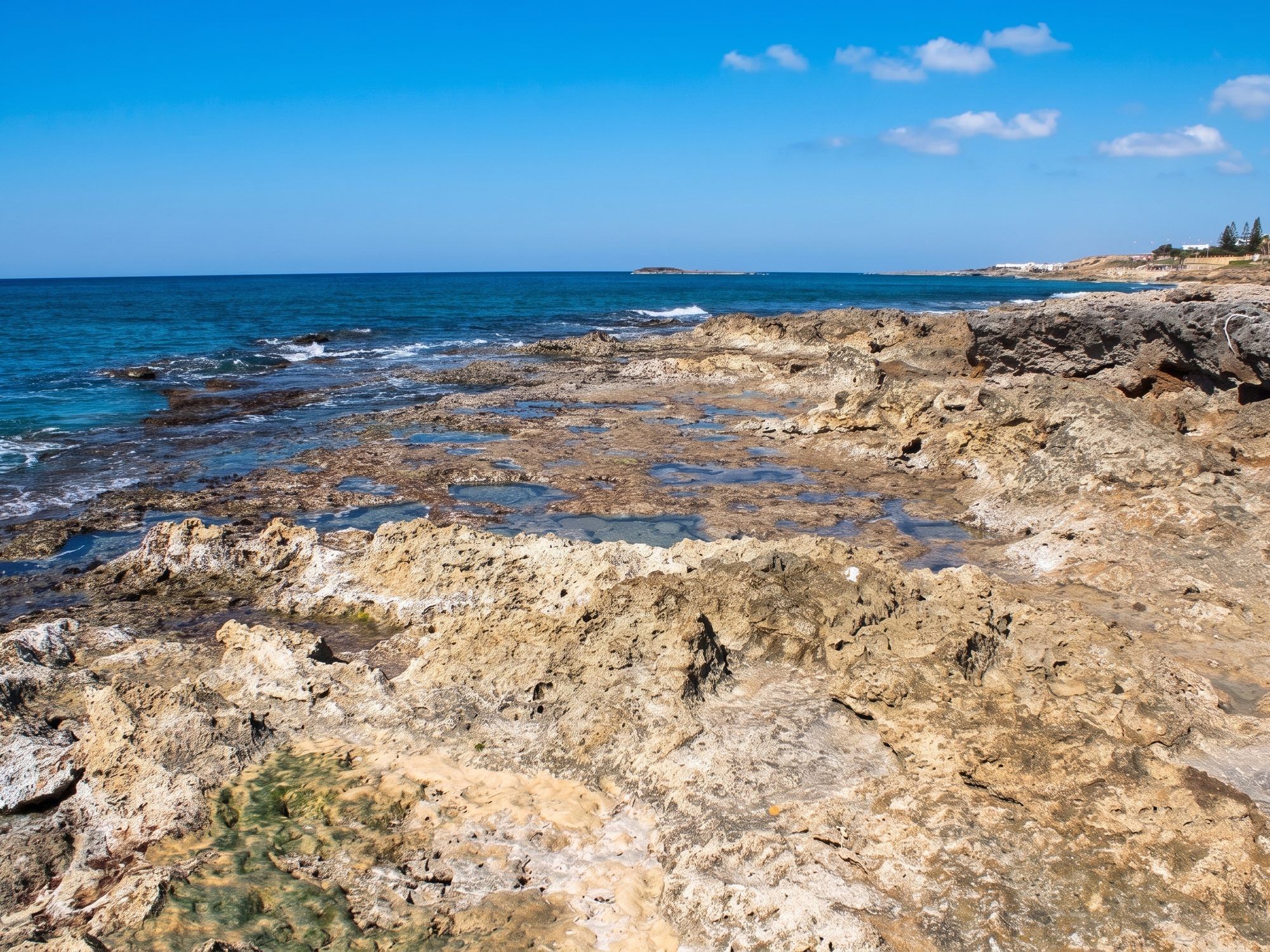 Scogli chiari e piatti sul mare blu a nord di Torre San Giovanni, con le case in lontananza