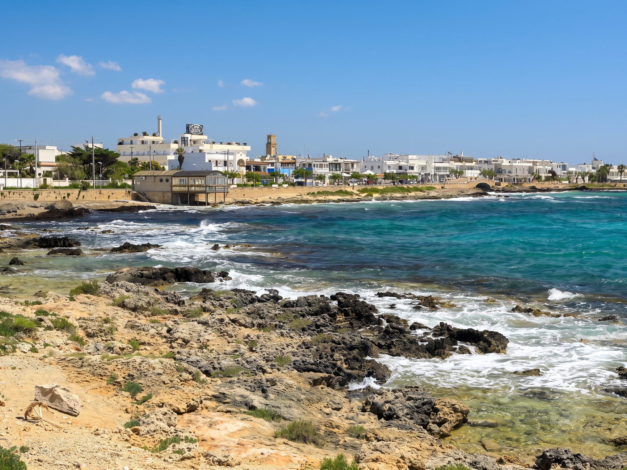 Scogli bassi e acqua turchese a nord di Torre San Giovanni, con il paese bianco sullo sfondo